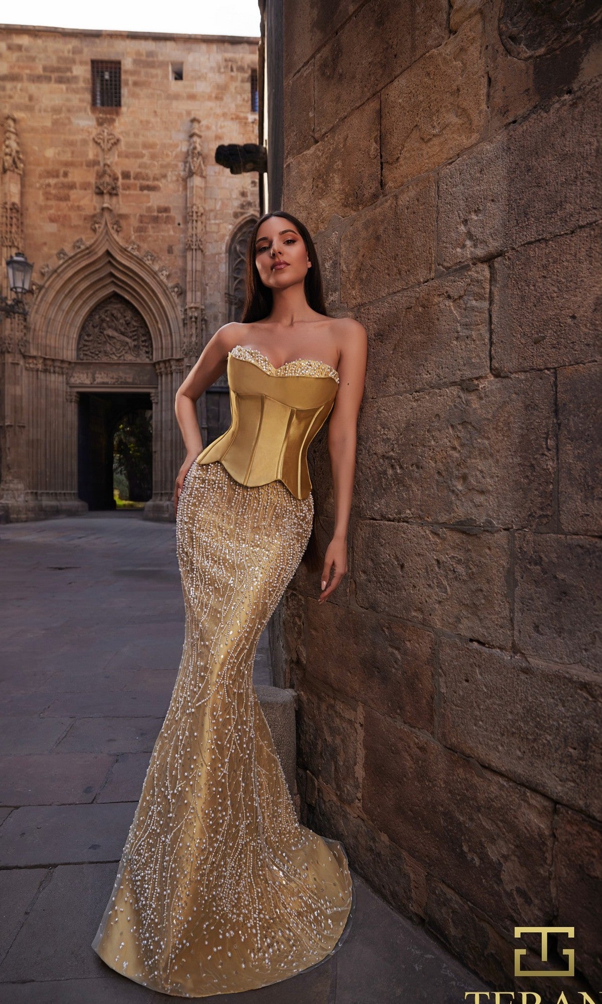 A woman in a gold Long Formal Dress (Terani 261E6238) stands against a stone wall in a historic courtyard.